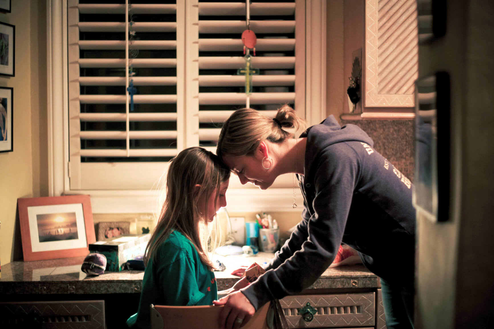 mother standing next to daughter sitting at desk with their foreheads pressed against each other 